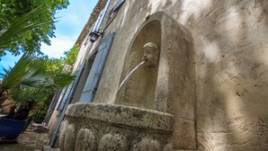 A stone fountain with a sculpted figure, set against a building with blue shutters and a tiled roof.