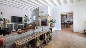 A spacious living room with a large sofa, a television, and a fireplace. There is a dining area in the background.