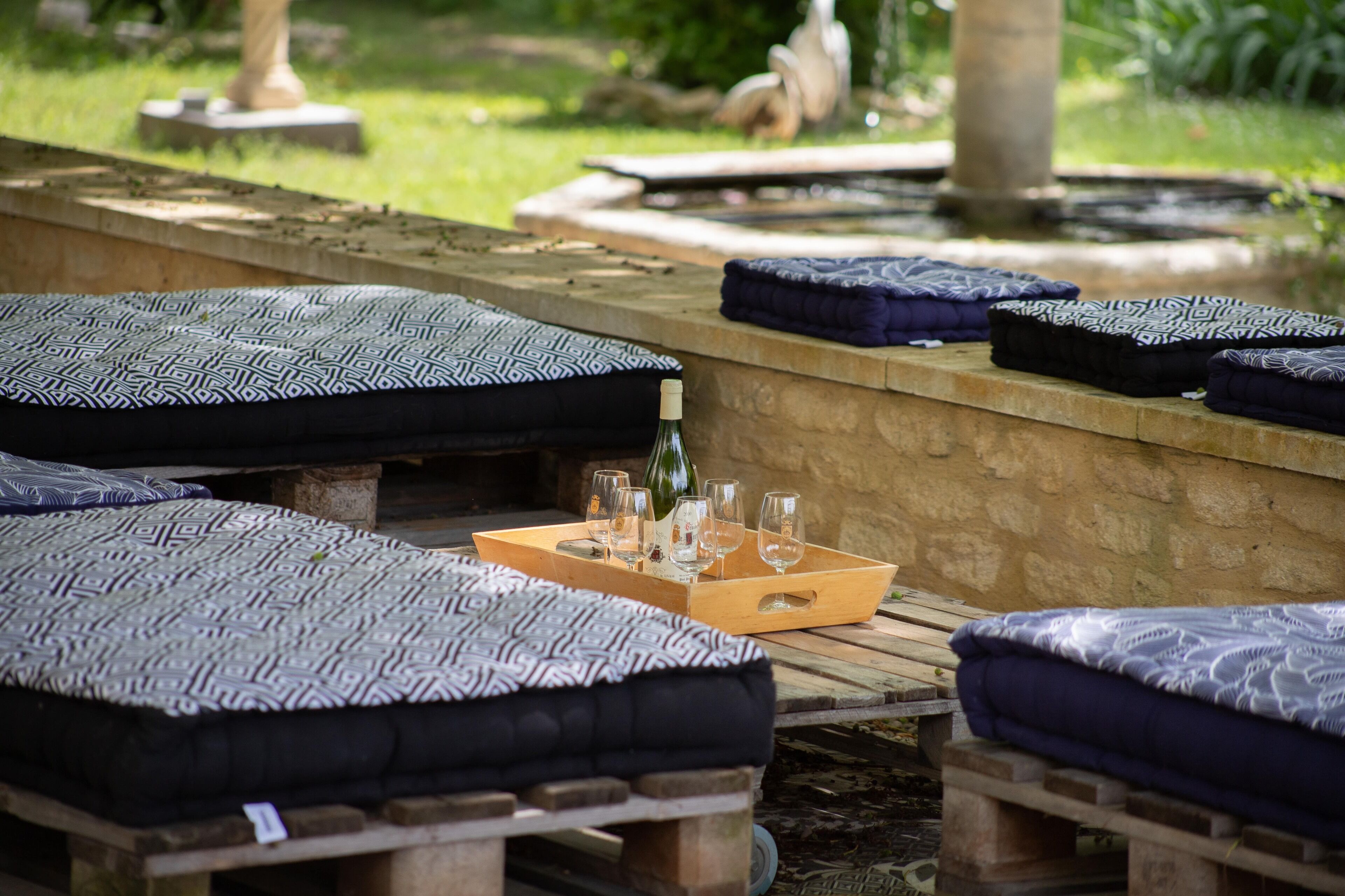 Mobiliario exterior con cojines estampados, una bandeja de madera con vasos y una botella, todo frente a un muro de piedra con un jardín de fondo.