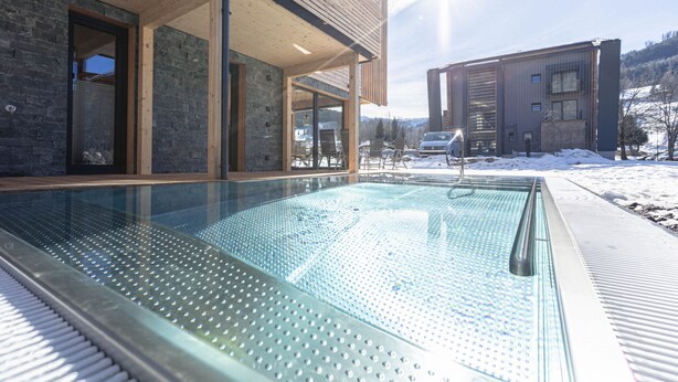 A modern building with a glass-enclosed pool area, wooden deck, and a view of a snowy landscape.