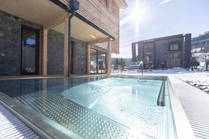 Un edificio moderno con zona de piscina cubierta de vidrio, terraza de madera y vistas a un paisaje nevado.