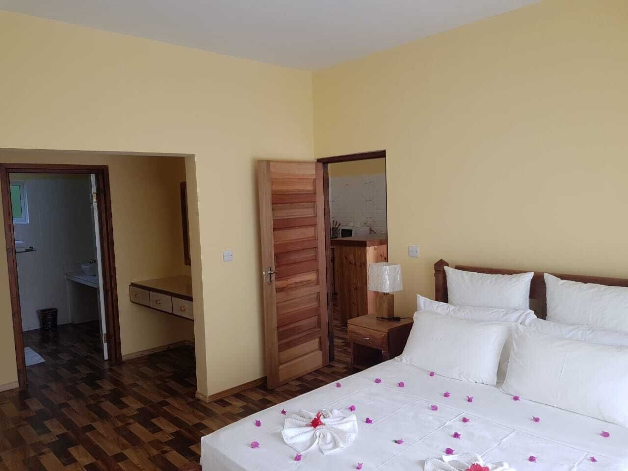 Een hotelkamer met een bed, een houten deur, een nachtkastje, een spiegel en een zichtbare badkamer door een geopende deur.