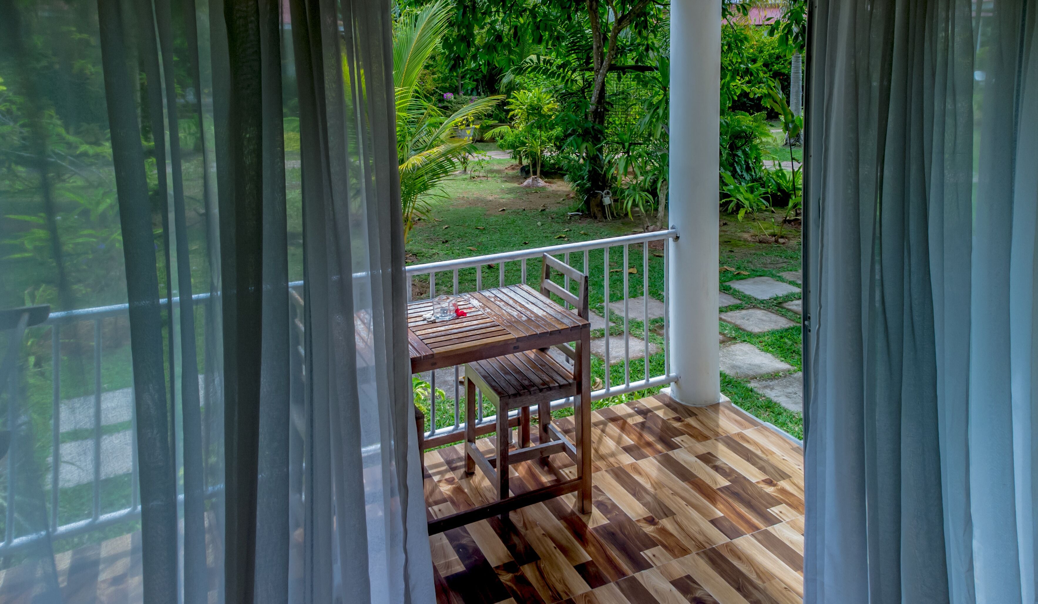 Una terraza de madera con una mesa y una silla, rodeada por una barandilla blanca, con vistas a un jardín con diversas plantas y un sendero de piedra.