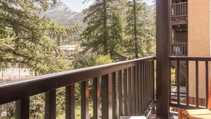 Ein Balkon mit Holzgeländer, der auf eine gebirgige Landschaft mit dichten Wäldern und einem Gebäude im Hintergrund blickt.