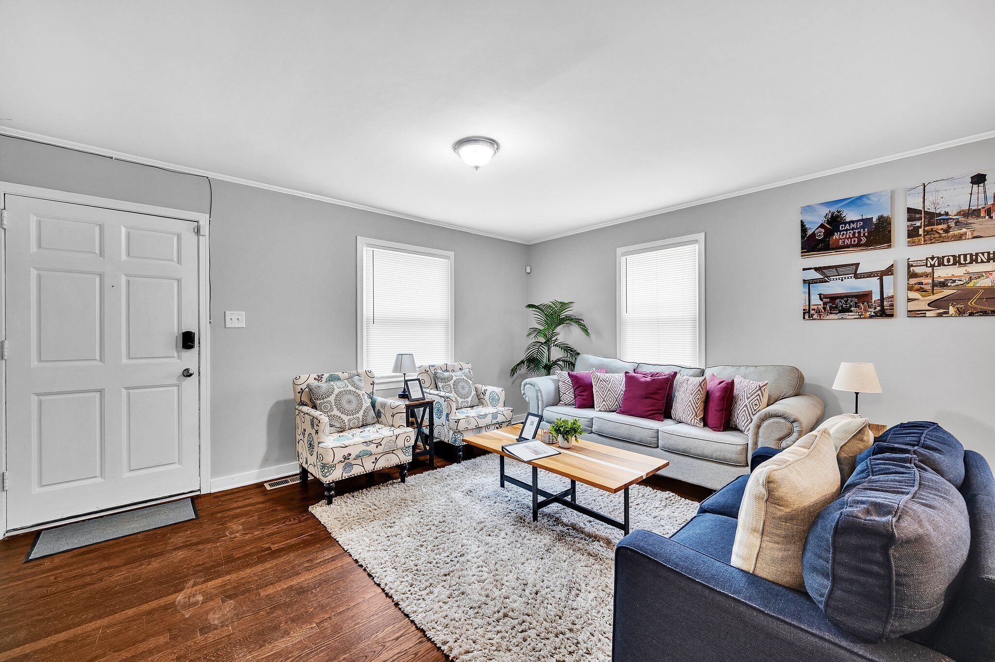 A living room with a sofa, two armchairs, a coffee table, and a rug. There are two windows with blinds, a door, and a ceiling light.
