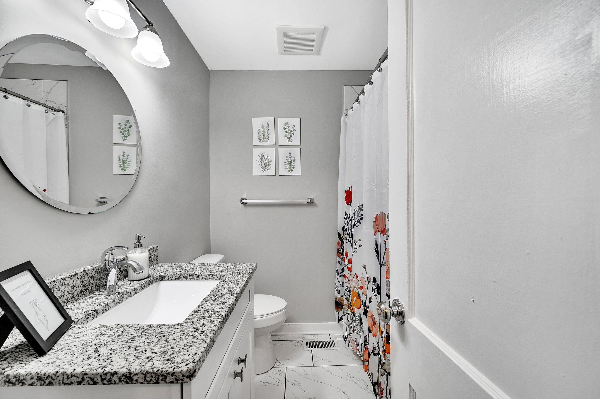 Een badkamer met een wastafel met granieten blad, een wit toilet, een spiegel met verlichting en een douchegordijn met bloemenmotieven.