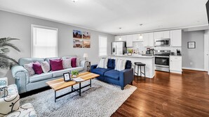 Un salón moderno con un sofá gris, un sofá azul y una mesa de centro de madera. Hay una zona de cocina con armarios blancos y electrodomésticos de acero inoxidable.