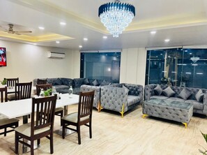 A modern living room with a dining area, grey sofas, wooden chairs, a glass coffee table, and a chandelier. - Hotel the Archer (Gurugram)