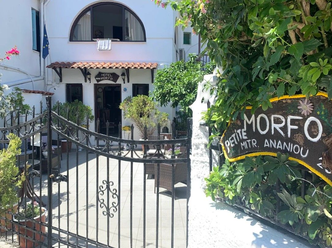 A hotel entrance with a wrought iron gate, a sign reading 'MORFO PETITE MTR. ANANIOU,' and a white building with a tiled roof.
