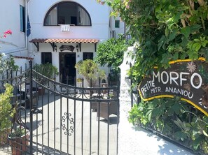 Una entrada de hotel con una reja de hierro forjado, un cartel que dice 'MORFO PETITE MTR. ANANIOU' y un edificio blanco con techo de tejas.