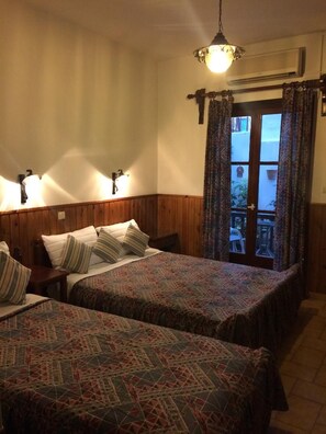 A room with two beds, a wooden headboard, a hanging light fixture, and a view through a door to an outdoor area. - Pettite Morfo (Skiathos)