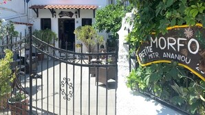 Una entrada de hotel con una reja de hierro forjado, un cartel que dice 'MORFO PETITE MTR. ANANIOU' y un edificio blanco con techo de tejas.