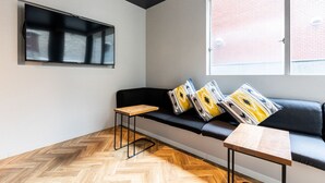 A modern interior with a black sofa, two wooden side tables, a mounted TV, and a window with a view of a brick building. - Akasaka The Hostel (Tokyo)