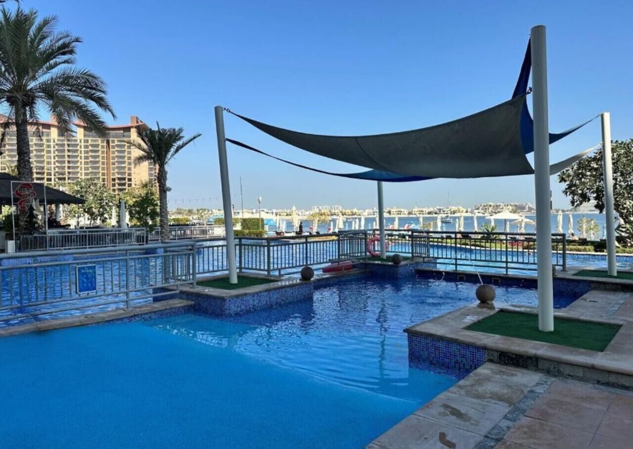 A swimming pool with a shaded area, surrounded by a fence, and palm trees in the background.