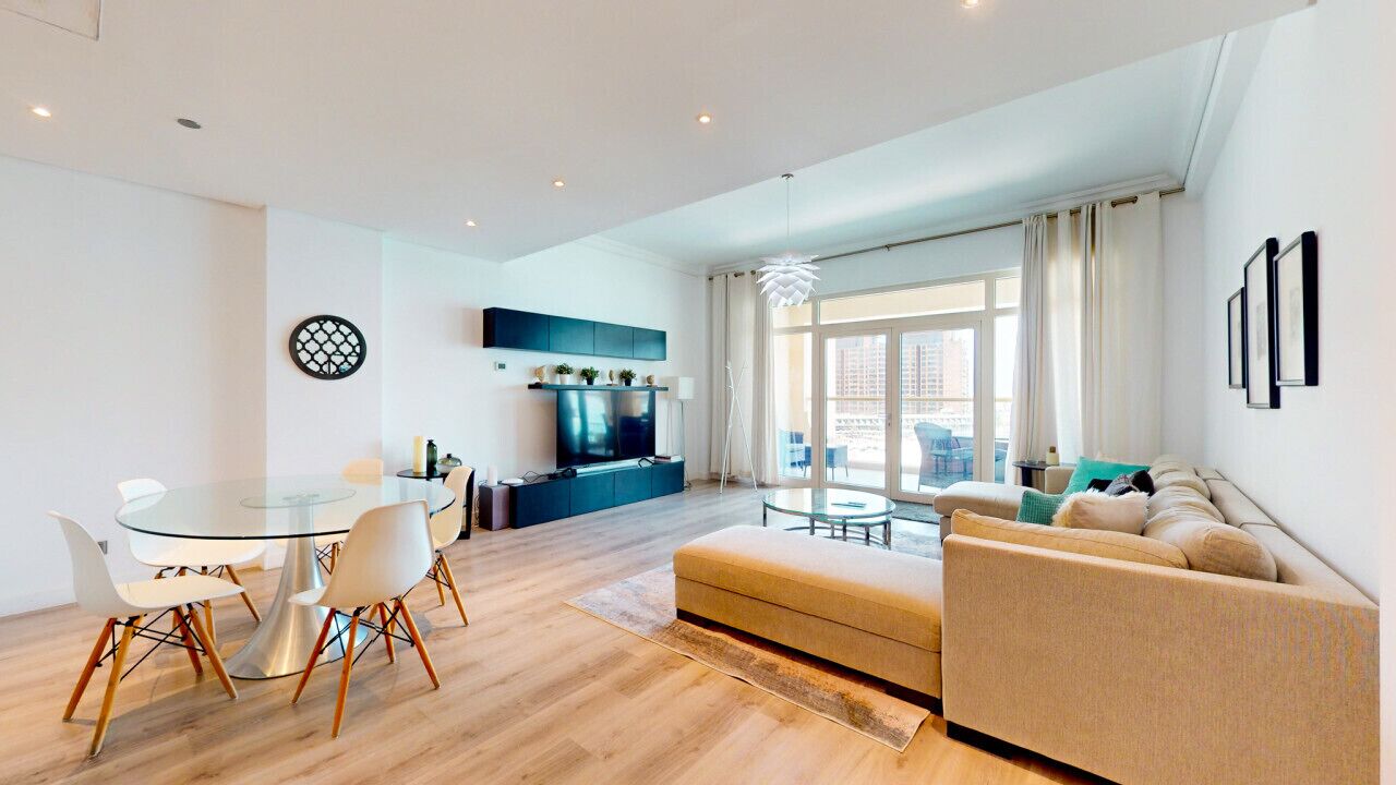A modern living room with a glass dining table, beige sofa, and a TV mounted on the wall.