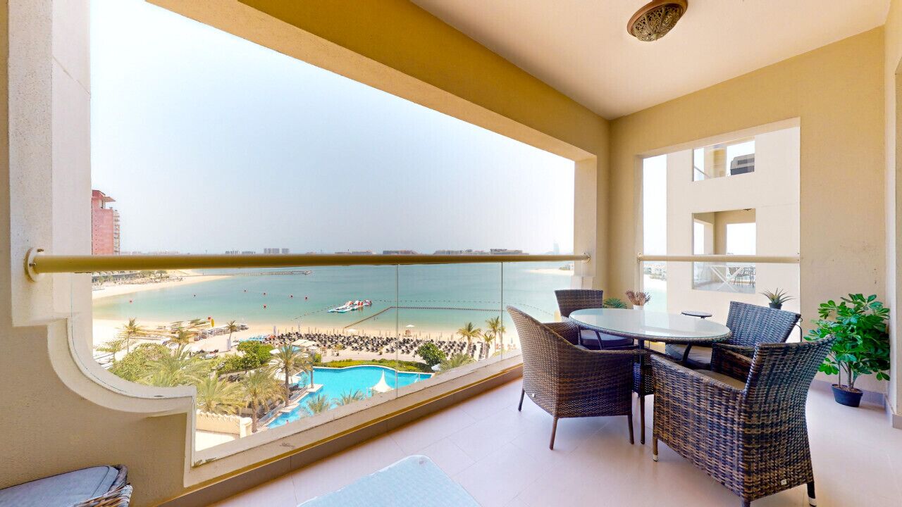 A balcony with a view of a beach and pool, wicker furniture, and a potted plant.