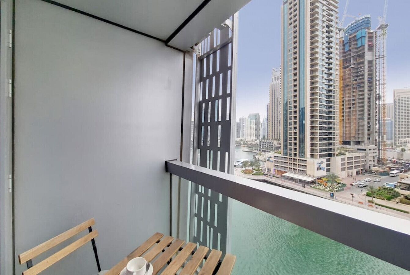 Un balcon avec un banc en bois et une table, d’où l’on peut admirer un paysage urbain avec de hauts immeubles et une étendue d’eau.