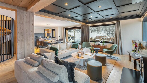 Ein modernes Wohnzimmer mit einer großen Couch, einem Couchtisch und einem Fernseher. Ein Essbereich mit Tisch und Stühlen ist ebenfalls vorhanden. Große Fenster bieten einen Blick ins Freie.
