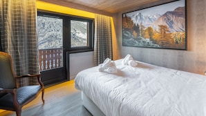 Ein Schlafzimmer mit einem großen Bett, Holzboden, einem fenster mit Vorhängen, einem Sessel und einem Gemälde einer Berglandschaft an der Wand.