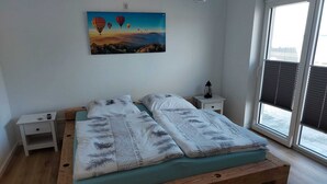 Quarto com cama de madeira, mesas de cabeceira brancas, pintura de balões de ar quente e vista pela janela.