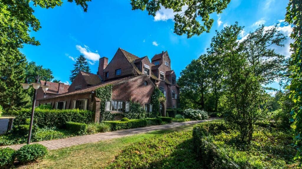A large, multi-story brick house with a prominent gabled roof, surrounded by lush greenery and a well-maintained garden.
