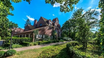 A large, multi-story brick house with a prominent gabled roof, surrounded by lush greenery and a well-maintained garden.