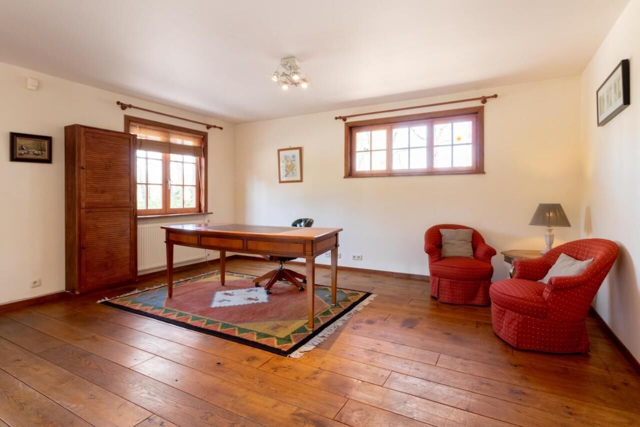 Una habitación con suelo de madera, una mesa de madera, dos sillones rojos, una lámpara y dos ventanas con marcos de madera.