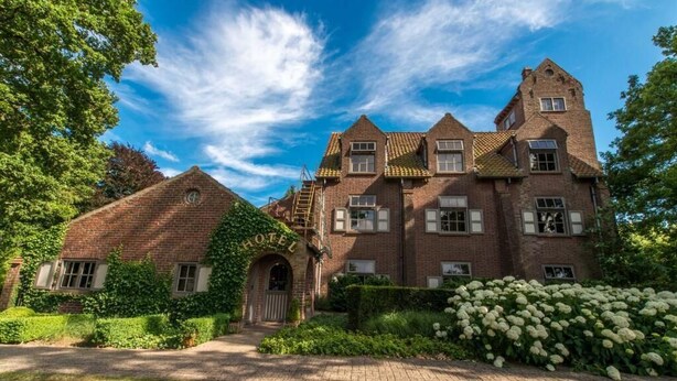Ein großes Backsteingebäude mit einem zentralen Turm, zahlreichen Fenstern und einem gepflegten Garten mit weißen Blüten.