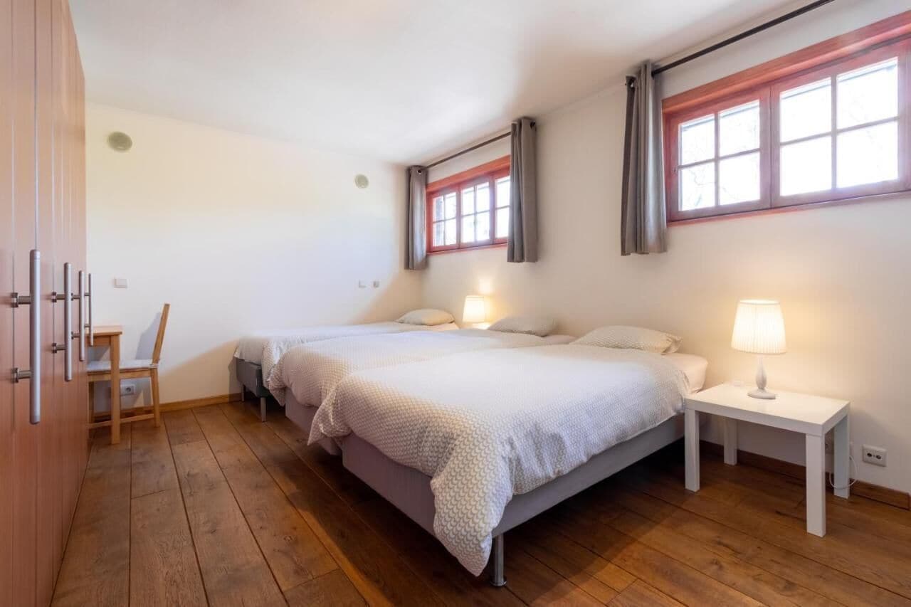 Un dormitorio con dos camas, piso de madera, una silla y una mesita de noche con lámpara.