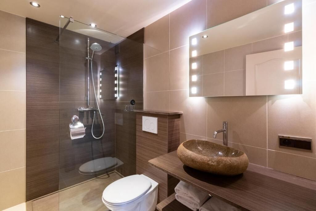 A modern bathroom with a glass-enclosed shower, a white toilet, a wooden vanity with a large stone sink, and a mirror with lights.