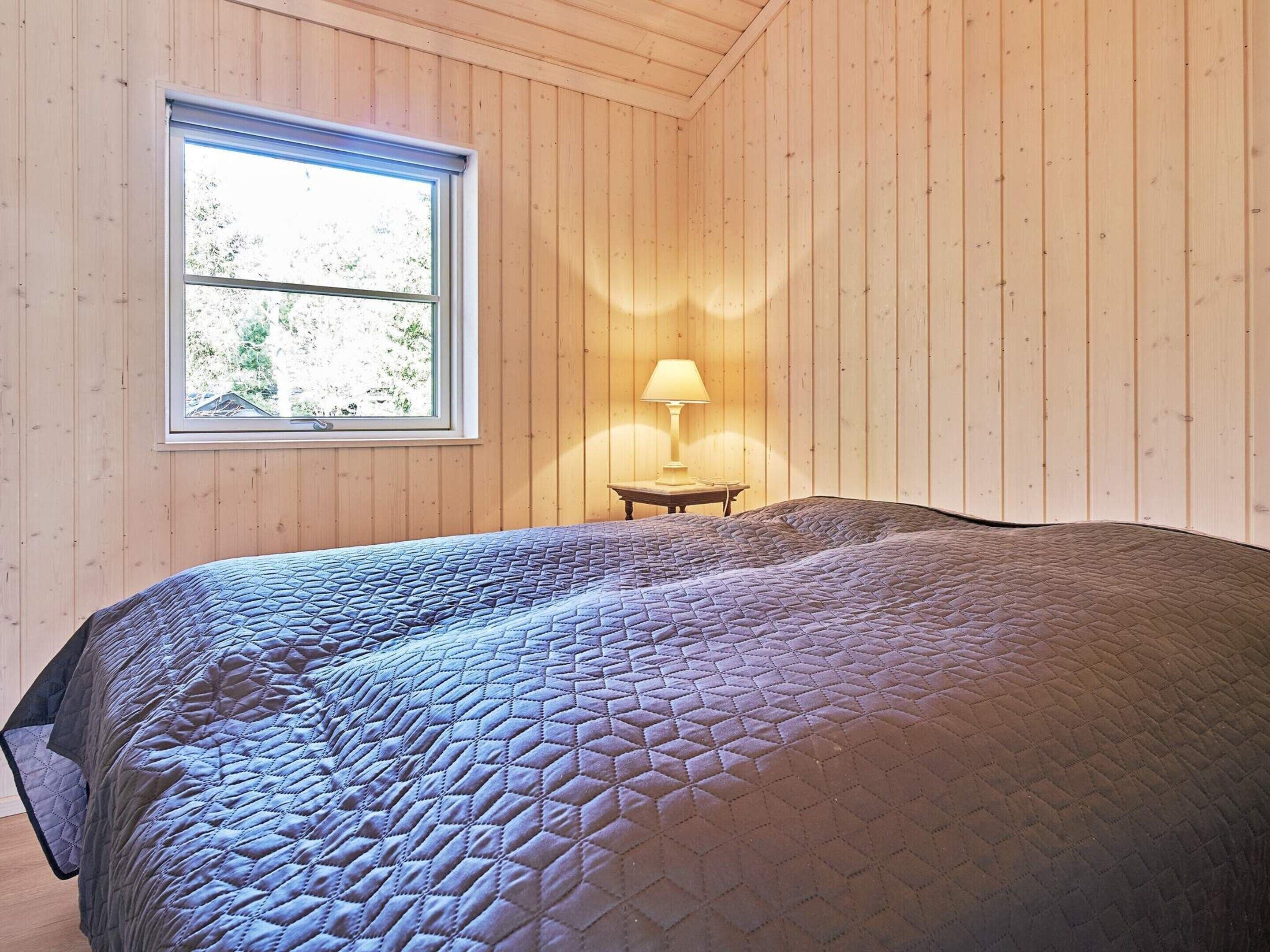 Ein Zimmer mit Holzwänden, einem Bett mit gesteppter Decke, einem Fenster und einer Nachttischlampe.