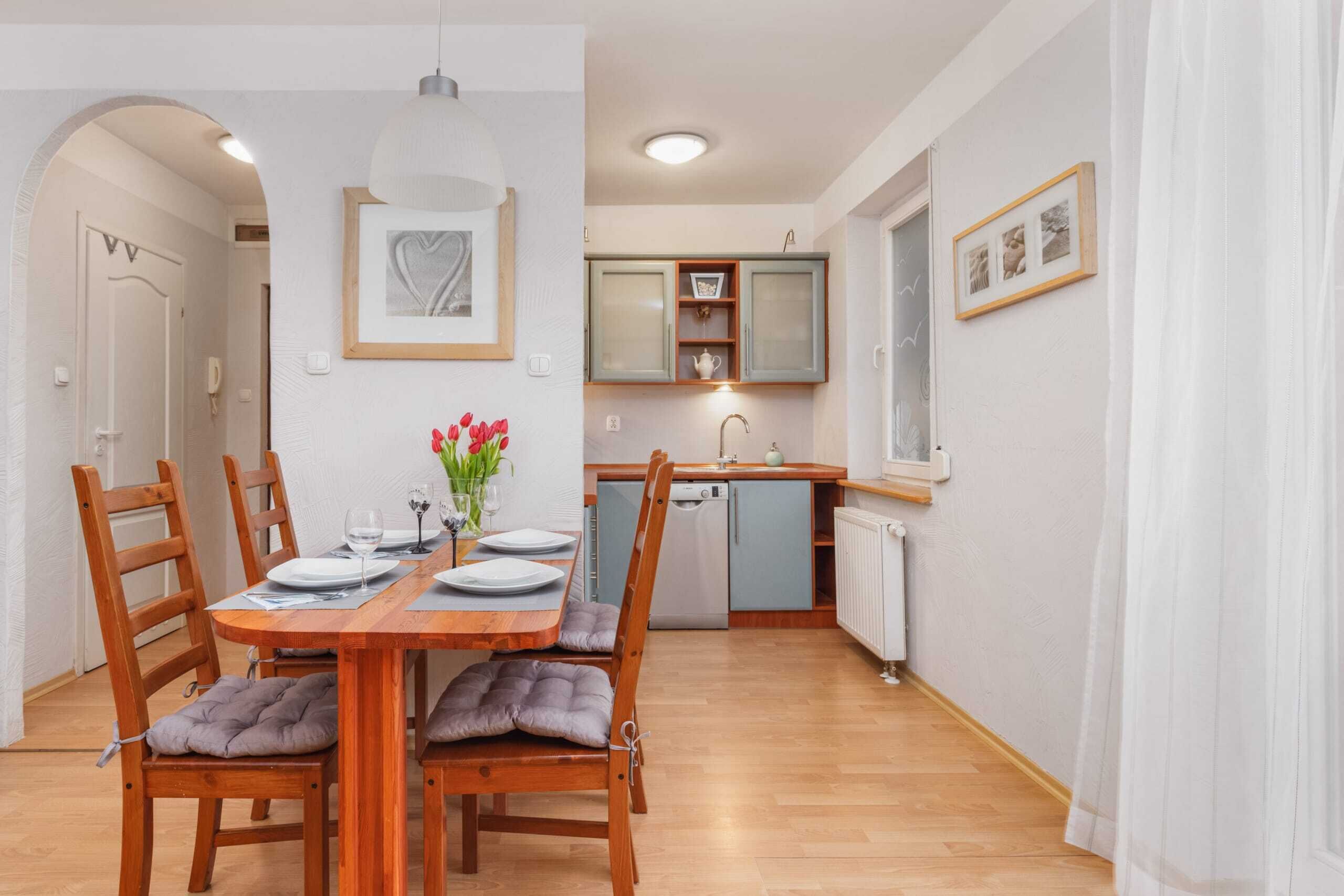 Een eetgedeelte met een houten tafel voor vier personen, beklede stoelen en een bloevas. Achtergrond toont een keuken met een vaatwasser en een raam.