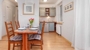 Un comedor con mesa de madera preparada para cuatro personas, sillas acolchadas y un florero. Se ve una cocina con lavavajillas y ventana al fondo.