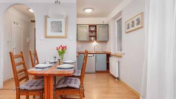 Een eetgedeelte met een houten tafel voor vier personen, beklede stoelen en een bloevas. Achtergrond toont een keuken met een vaatwasser en een raam.