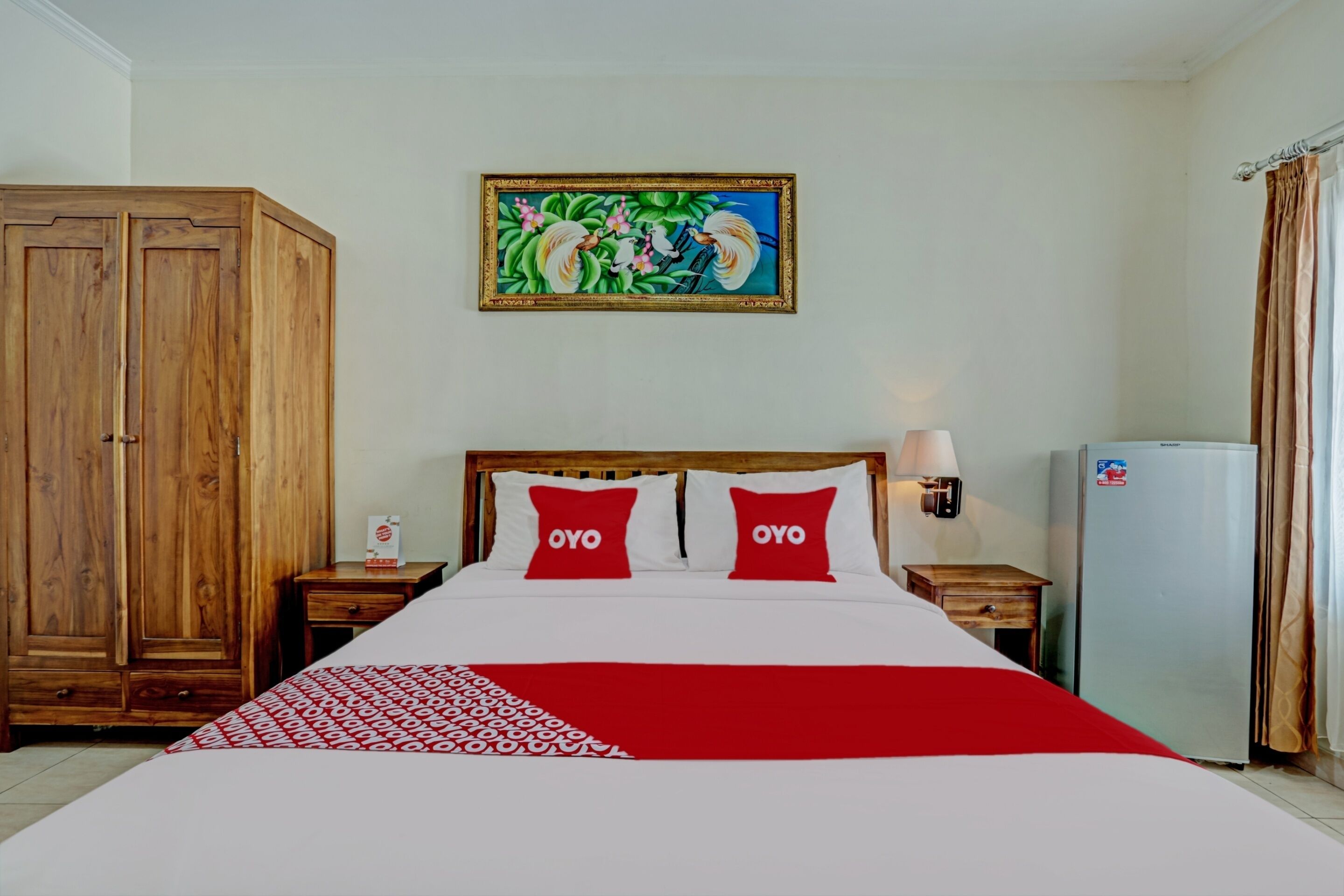 A hotel room with a large bed, two red pillows, a wooden wardrobe, a wall-mounted painting, a bedside table with a lamp, and a refrigerator.