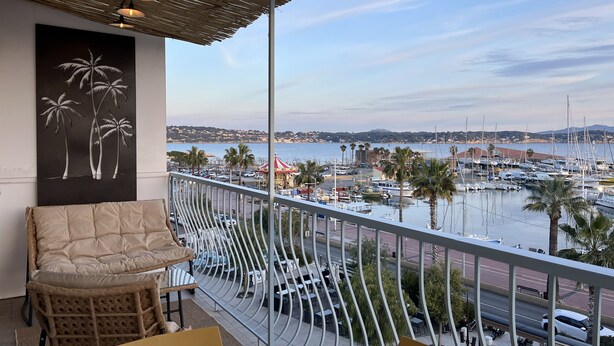 A balcony with wicker furniture, a view of a marina, and a painting of palm trees. - Panoramic sea view of port and beaches (Bandol)