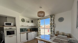 A compact kitchen with white cabinets, a microwave, an oven, and a sink. There is a dining table with chairs and a view of buildings through a window with patterned curtains. - Panoramic sea view of port and beaches (Bandol)