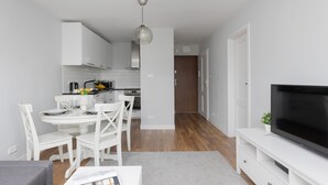 Une cuisine moderne avec des meubles blancs, un coin repas avec des chaises blanches, un canapé gris et un téléviseur à écran plat posé sur un meuble blanc.