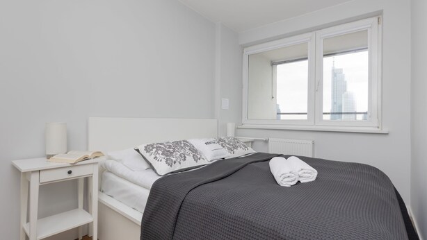 Une chambre à coucher bien rangée, avec un lit, une table de chevet dotée d’une lampe et une fenêtre donnant sur des immeubles.