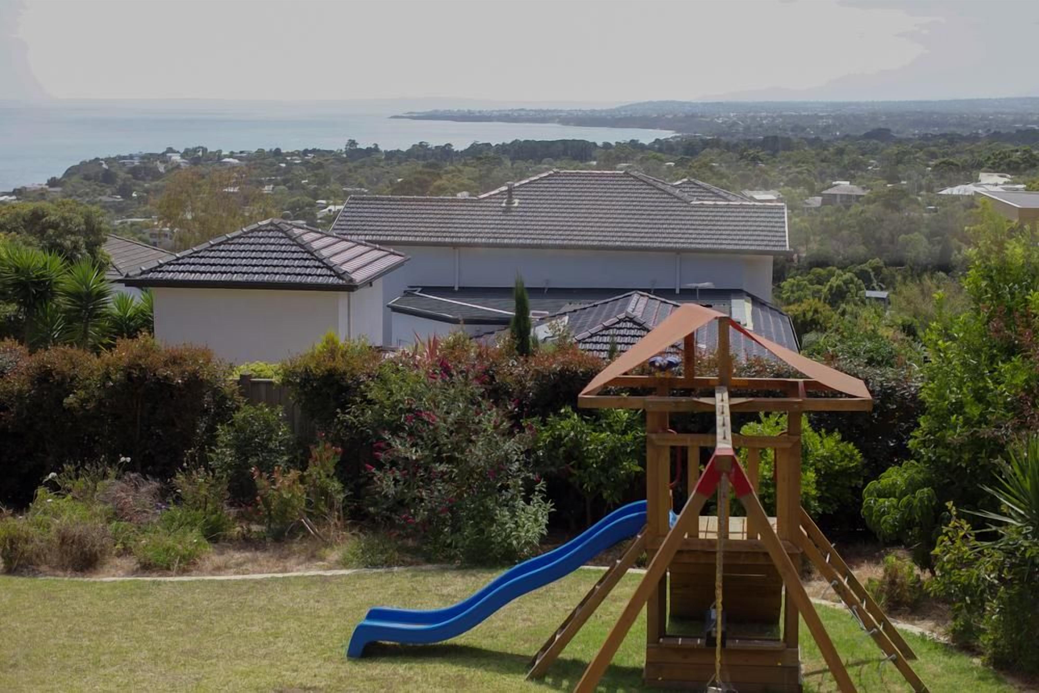 A residential area with a house, a wooden play structure with a slide, and a view of the sea.