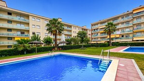 A swimming pool with a red border, surrounded by a well-maintained lawn and palm trees. - Apt, 4 pax LLoret de Mar - Fenals - wifi, community pool a few meters from the beach. Vivalidays Elia. (Lloret de Mar)