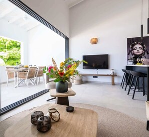Un salon moderne doté d’un grand téléviseur, d’un coin repas avec des chaises en osier et d’une table basse en bois agrémentée d’objets décoratifs.