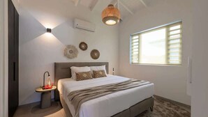 A bedroom with a bed, bedside table, wall decor, and a window with blinds. - Penthouse Mesa Bonita – Modern Caribbean Luxury (Jan Thiel)