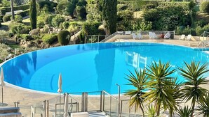 Una piscina con sillas de descanso y sombrillas, rodeada de vegetación y un edificio al fondo.