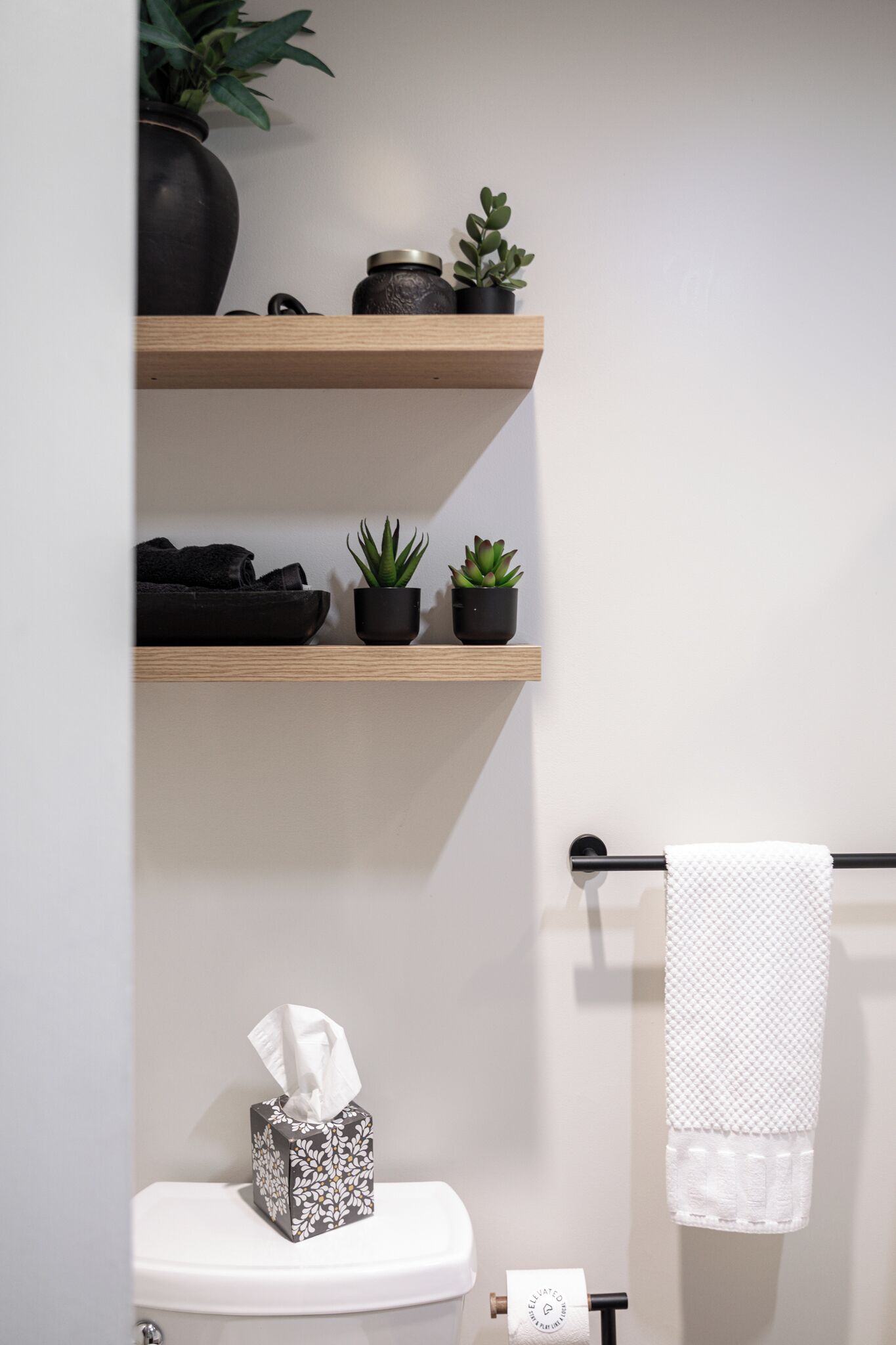Une salle de bain avec des toilettes, un porte-serviettes avec une serviette blanche, et deux étagères avec des plantes et un r écipient noir.