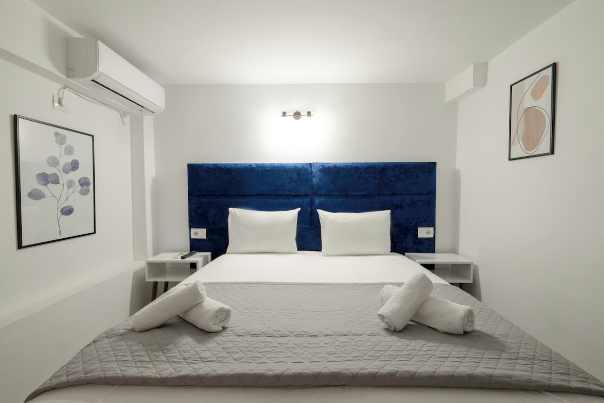 Habitación de hotel con una cama grande, cabecera azul, ropa de cama blanca, dos mesitas de noche y aire acondicionado instalado en la pared.