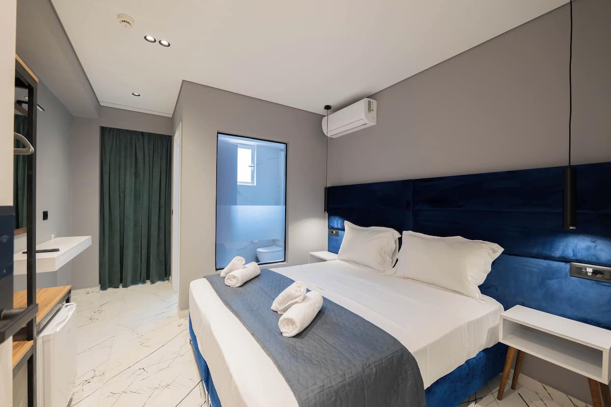 Una habitación de hotel moderna con una cama grande, una mesita de noche, un estante empotrado, una ventana con cortinas y un baño visible a través de la ventana.