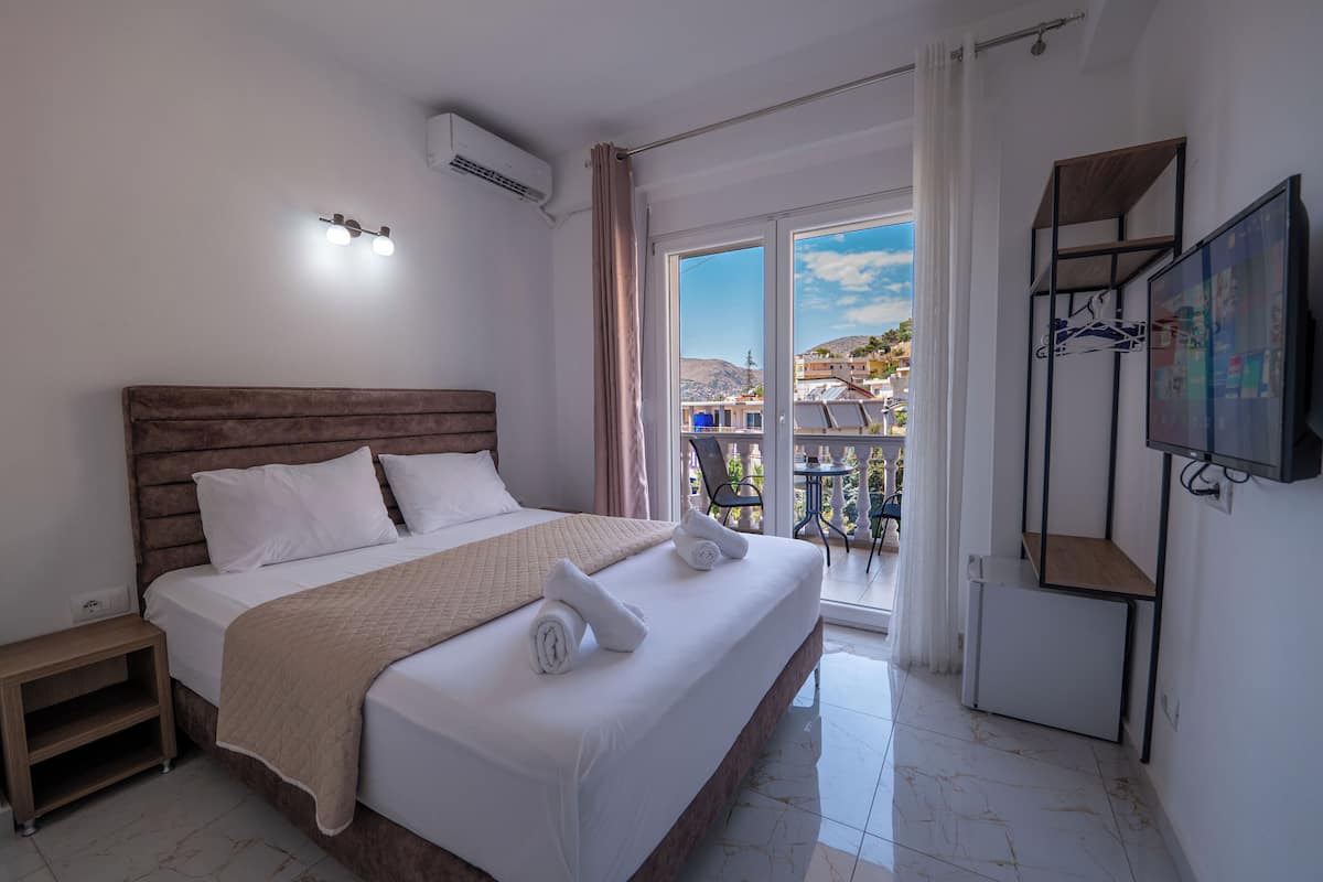 Habitación de hotel con cama, televisor, balcón y vista a edificios.