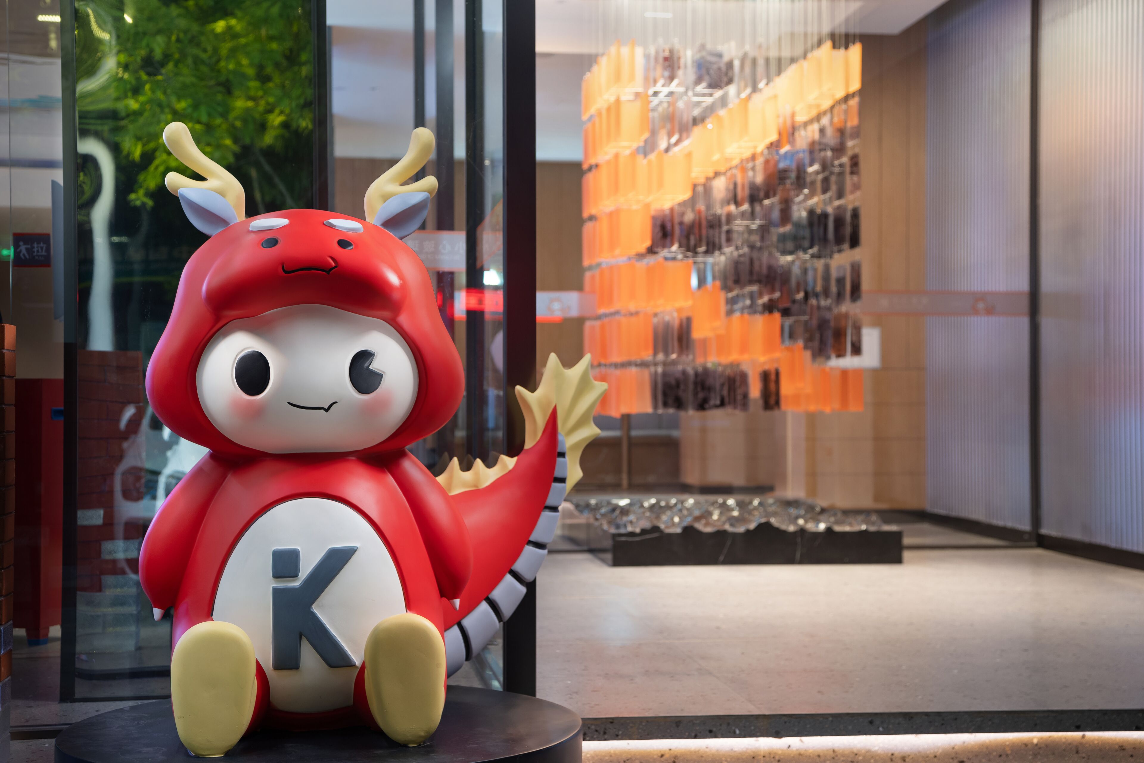 Un peluche rojo con una 'K' en su vientre, sobre una plataforma negra circular en un ambiente interior moderno con paredes de vidrio y una exhibición de objetos al fondo.