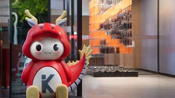 Un peluche rojo con una 'K' en su vientre, sobre una plataforma negra circular en un ambiente interior moderno con paredes de vidrio y una exhibición de objetos al fondo.